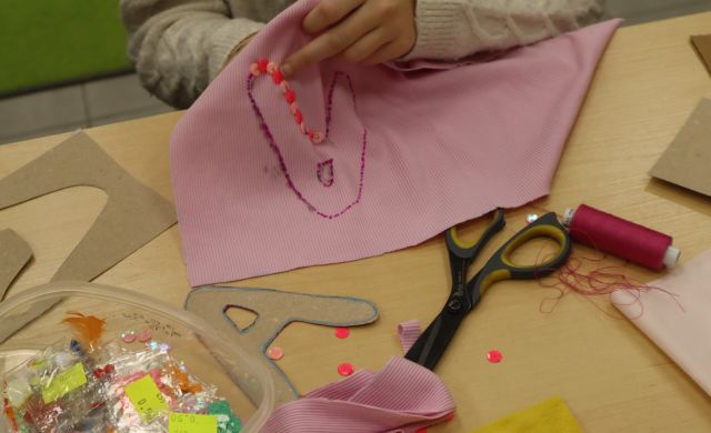 Na zdjęciu znajdują się dłonie dziewczynki, która za pomocą koralików wycina literkę A na różowym materiale, przed dziewczynką leżą nitki, koraliki, nożyczki oraz wycięte z kartoniku A. 