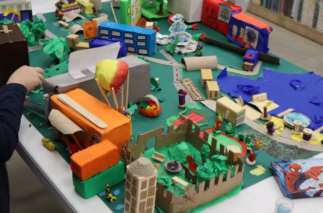 Na zdjęciu znajduje się makieta przedstawiająca miasto z budynkami, jeziorem, balonem drogami, roślinnością. Makieta znajduje się na stole.