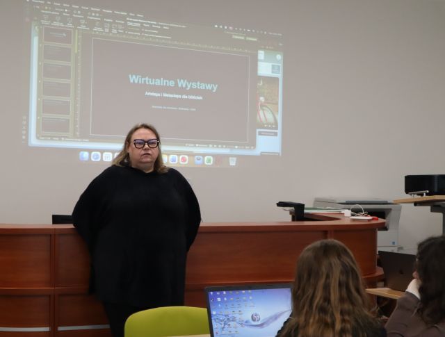 Na zdjęciu znajduje się kobieta, która opiera się o biurko, a za nią na ścianie wyświetlana jest prezentacja o Wirtualnej Wystawie. Przed kobietą znajdują się dwie dziewczyny przy stoliku z laptopem.