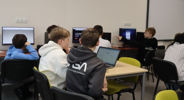 Na zdjęciu znajduje się młodzież przy stolikach z laptopami i przy komputerach i tworzą swoją wirtualną wystawę. Zajęcia odbywają się w sali komputerowej.