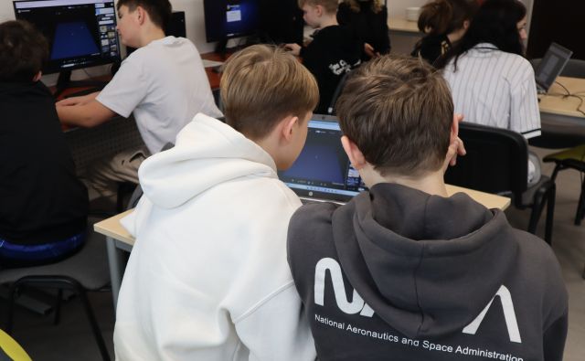 Na zdjęciu w centrum znajduje sie dwójka chłopców przy laptopie, w tle znajduje sie reszta uczniow przy stolikach z laptopami i przy komputerach. Zdjęcie zostało wykonane od tyłu.