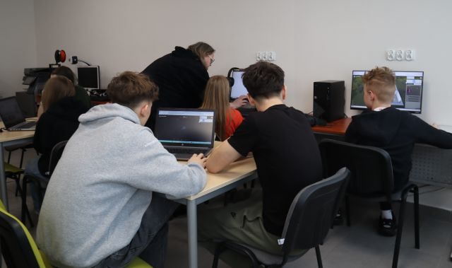 Na zdjęciu znajduje się dwójka chłopaków przy laptopie, za nimi siedzi chłopiec i dziewczynka przy komputerach, a przy dziewczynie nachyla się kobieta prowadząca warsztaty. Zdjęcie zostało wykonane od tyłu. Warsztaty odbywają się w sali komputerowej.