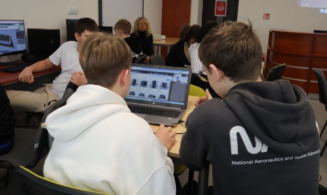 Na zdjęciu w centrum znajduje się dwójka chłopaków, którzy robią wirtualną galerię na laptopie. W tle znajduje się reszta uczestników przy laptopach. Warsztaty odbywają się w sali komputerowej. Zdjęcie zostało wykonane od tyłu.