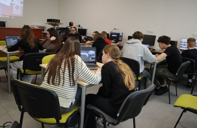 Na zdjęciu znajduje się młodzież przy stolikach z laptopami oraz przy komputerach i tworzą swoją wirtualną galerię. Warsztaty odbywają się w sali komputerowej.