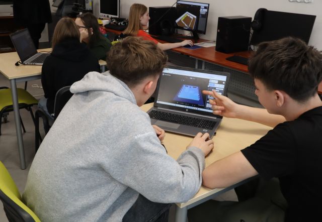 Na zdjęciu znajduje się dwójka nastolatków przy stoliku z laptopem, jeden z nich wskazuje na ekran przedstawiający budynek palcem. W tle znajdują się dwie dziewczyny przy stoliku z laptopem i jedna przy komputerze. Warsztaty odbywają się w sali komputerowej.