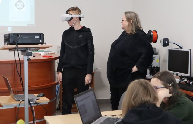Na zdjęciu znajduje się chłopak w okularach VR, obok niego po prawej stronie stoi kobieta, po lewej stronie znajduje się rzutnik, a przed nimi znajdują się dwie dziewczyny przy stoliku z laptopem. Warsztaty odbywają się w sali komputerowej.