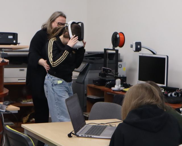 Na zdjęciu znajduje się dziewczyna w okularach VR, za nią stoi kobieta i trzyma ją delikatnie w pasie. Przed nimi przy stoliku z laptopem siedzą dwie kobiety. Warsztaty odbywają się w sali komputerowej.