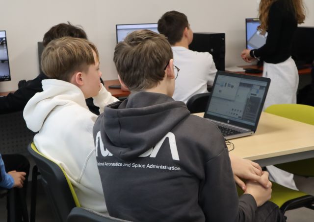 Na zdjęciu znajduje się dwójka chłopców przy stoliku z laptopem, na którym tworzą swoją wirtualną galerię, w tle znajdują się chłopcy przy komputerach. Warsztaty odbywają się w sali komputerowej. 