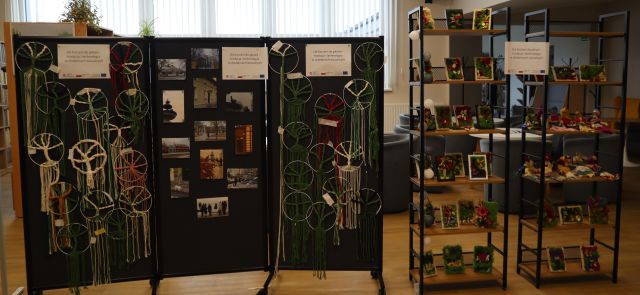 Na zdjęciu znajduje się potrójna czarna tablica, na której powieszone są drzewka szczęścia na kole, a po środku zdjęcia. Po prawej stronie zdjęcia znajdują się dwie szafki z półkami, na których stoją obrazy z mchu. W tle znajdują się okna.