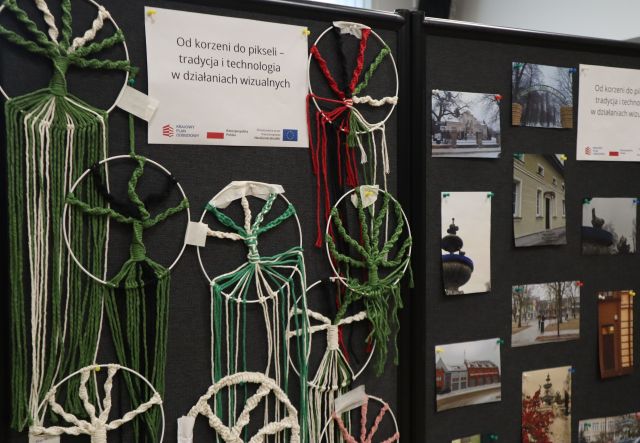 Na zdjęciu znajdują się powieszone na czarnej tablicy drzewa szczęścia na kole i zdjęcia po prawej stronie. Do góry powieszona jest kartka z nazwą projektu. Zdjęcie zostało wykonane od boku.