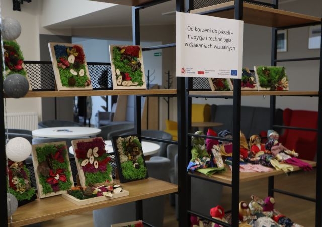 Na zdjęciu znajdują się półki, na których stoją obrazy z mchu. Po środku wisi kartka z nazwą projektu.