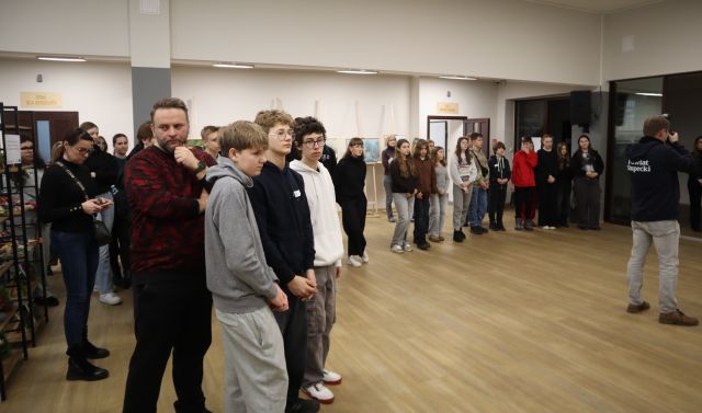 Na zdjęciu znajduje się młodzież stojąca w półkolu na holu biblioteki. Po prawej stronie zdjęcia przed młodzieżą stoi mężczyzna z aparatem.