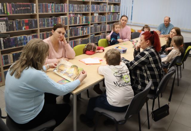Na zdjęciu znajdują się dzieci wraz z rodzicami przy złączonych stołach, na brzegu siedzi kobieta i czyta książkę. W tle znajdują się półki z książkami.