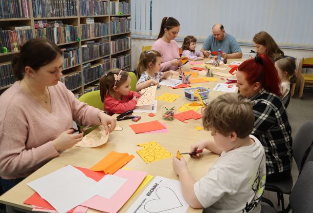 Na zdjęciu zmnajdują się dzieci wraz z rodzicami przy złączonych stołach, na których leżą kolorowe kartki, kleje i nożyczki. W tle znajdują się okna oraz półki z książkami.
