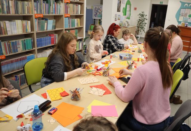 Na zdjęciu znajdują się cztery kobiety wraz z dziećmi przy złączonych stołach i wycinają serduszka z kolorowych kartek znajdujących sie na stole wraz z nożyczkami, klejem i kredkami. W tle znajdują się półki z książkami.
