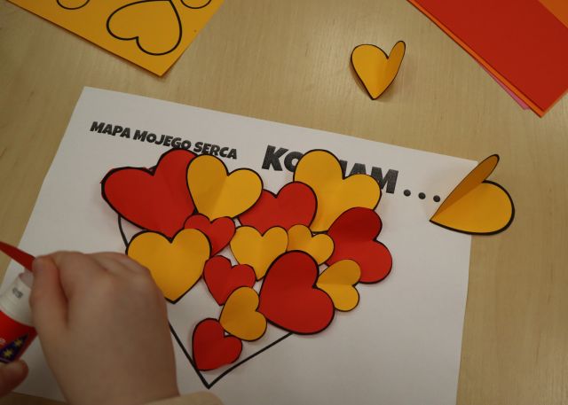 Na zdjęciu znajduje się biała kartka z przyklejonymi pomarańczowymi i czerwonymi serduszkami w serduszku, na kartce nad sercem znajduje się napis Kocham. Na zdjęciu znajdują się również rączki dziecka, które klei kolejne serduszko.
