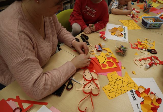 Na zdjęciu znajduje się kobieta wraz córką, które wspólnie tworzą czerwony łańcuch z serduszek. Na stole przed nimi znajdują się kolorowe kartki, nożyczki. Zdjęcie zostało wykonane od boku.