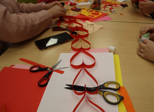 Na zdjęciu znajduje się stół, na którym leżą kolorowe kartki i nożyczki oraz w centrum czerwony łańcuch z serduszek. Przy stole znajdują się dłonie uczestników, którzy łańcuch.