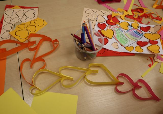 Na zdjęciu na stole wśród kolorowych kartek i kredek znajduje się kolorowy łańcuch z sercami.