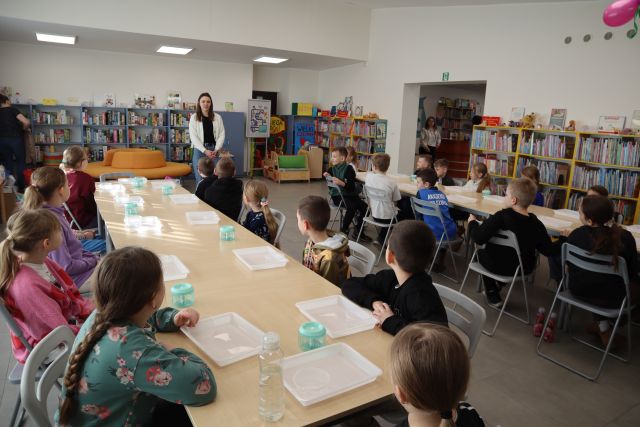 Na zdjęciu znajduje się dwadzieścia pięć dzieci przy złączonych stolikach w dwóch rzędach, pomiędzy stolikami stoi kobieta. W tle znajdują się półki z książkami.