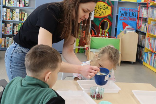 Na zdjęciu znajduje się dwójka dzieci przy stolikach i kobieta nachylająca się aby dodać jogurt do małego miksera znajdującego się na stoliku. W tle znajdują się półki z książkami.