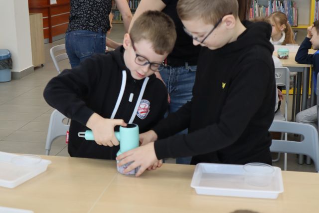 Na zdjęciu znajduje się dwójka chłopców przy stoliku i jeden pomaga drugiemu w miksowaniu malin z jogurtem na lody. W tle znajduje się reszta uczestników.