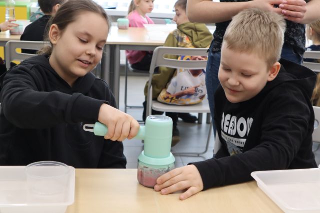 Na zdjęciu znajduje się chłopiec z dziewczynką i razem miksują owoce z jogurtem. W tle znajduje się reszta uczestników przy stolikach.