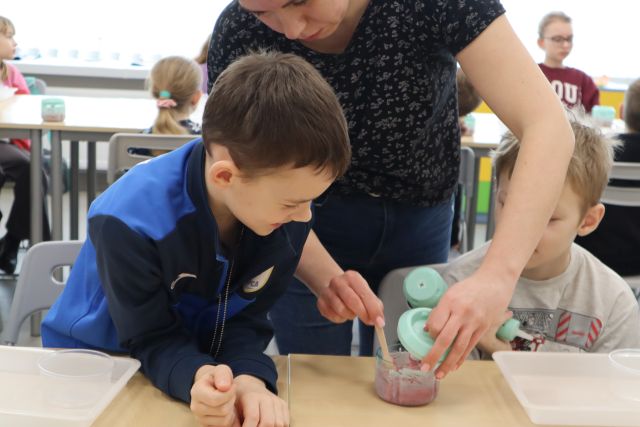 Na zdjęciu znajduje się dwójka chłopów przy stoliku oraz kobieta między nimi otwierająca mały mikser. W tle znajduje się reszta uczestników.