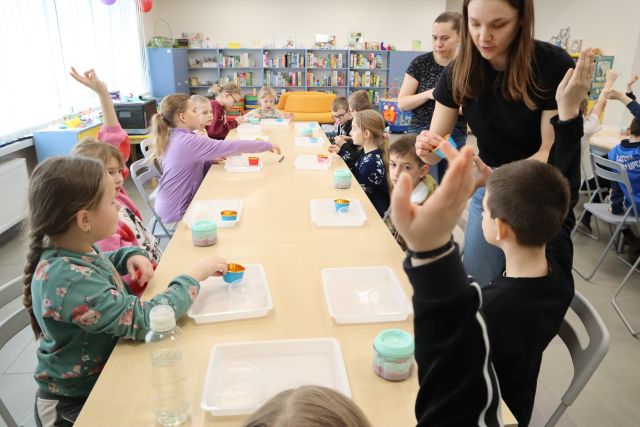Na zdjęciu znajdują się dzieci przy złączonych stolikach, część z nich ma podniesioną rękę, przed nimi znajdują się podstawki plastikowe i pudełka na lody, które rozdają dwie kobiety po prawej. W tle znajdują się półki z grami.