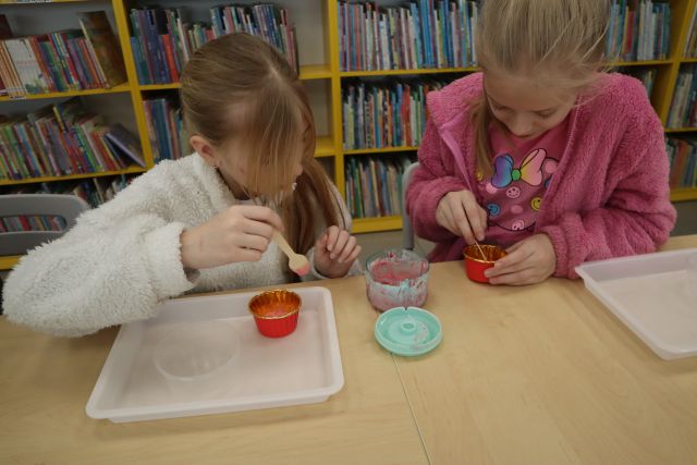 Na zdjęciu znajdują się dwie dziewczynki, które nakładają lody z miksera do pojemniczków. Na stoliku znajdują się również plastikowe podstawki. W tle znajdują się półki z książkami.