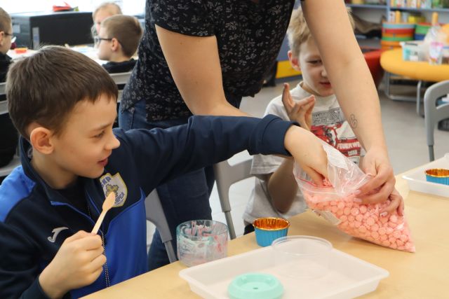 Na zdjęciu znajduje się dwóch chłopców przy stoliku, jeden z nich sięga po małe bezy, które trzyma kobieta za nim. Przed nimi na stole leżą przezroczyste pojemniczki. W tle znajduje się reszta uczestników przy stołach.
