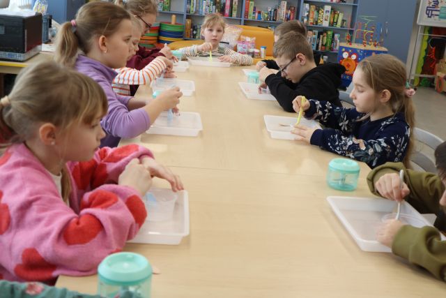 Na zdjęciu znajdują się dzieci przy złączonych stolikach, które mieszają masę w plastikowych pojemniczkach. W tle znajdują się półki z grami.