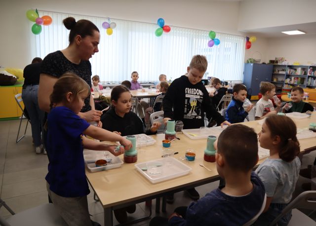 Na zdjęciu znajdują się dzieci przy złączonych w dwóch rzędach stolikach, przy których stoją dwie kobiety i pomagają w miksowaniu owoców. W tle znajdują się okna przyozdobione balonami.