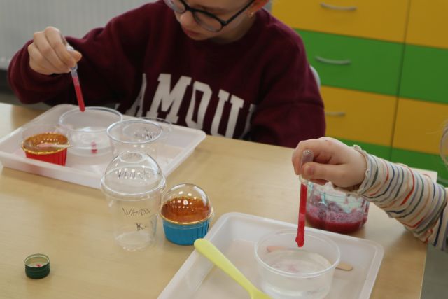 Na zdjęciu przy stoliku znajduje się dziewczynka, a po prawej stronie tylko ręka innej dziewczynki, obie za pomocą pipety wkraplają czerwoną ciecz do małych pojemniczków, które znajdują się na podstawkach przed nimi.