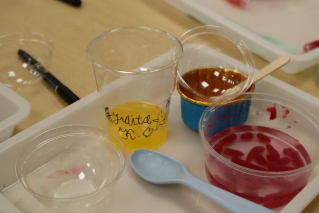 Na zdjęciu znajduje się plastikowa podstawka, na której stoi plastikowy kubeczek z podpisem oraz żółtym sokiem na dnie, obok stoi plastikowe zakrycie do kubka, okrągły pusty pojemniczek, pojemnik z wodą i czerwonymi żelkami w środku oraz niebieska łyżeczka obok.