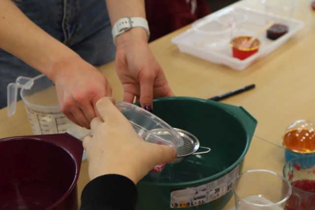 Na zdjęciu znajduje się stół, na którym stoi miska, nad którą jedna osoba trzyma małe sitko, a druga przelewa przez nie ciecz z plastikowego pojemnika. Na stole dookoła znajdują się kolorowe pojemniczki.