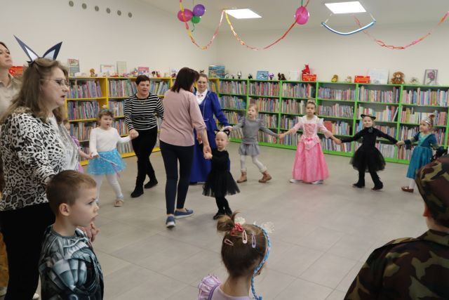 Na zdjęciu znajduje się grupa dzieci wraz z kobietami tańcząca w kółku, a w środku niego tańczy kobieta z dziewczynką. W tle znajdują się półki z książkami, a na suficie znajdują się girlandy i balony.