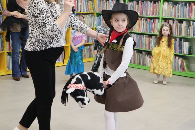 Na zdjęciu znajduje się dziewczynka ubrana za kowbojke, obok niej stoi kobieta, a za nimi znajdują się inni uczestnicy balu. W tle znajdują się półki z książkami.