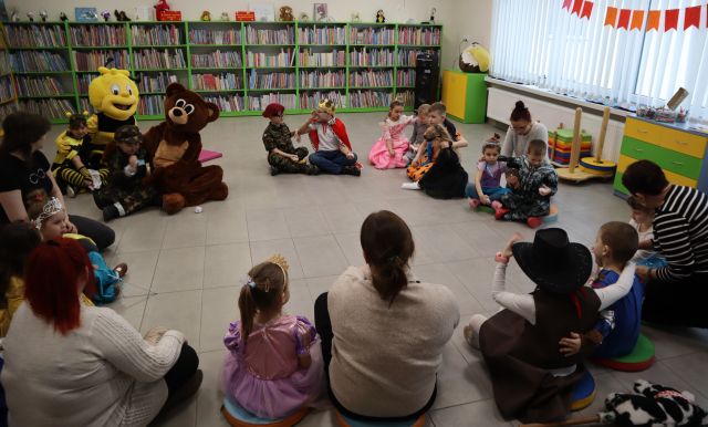 Na zdjęciu znajduje się grupa dzieci na ziemi wraz z misiem i pszczółką. W tle znajdują się półki z książkami, głośnik oraz okna po prawej stronie.
