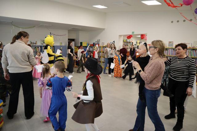Na zdjęciu znajduje się grupa dzieci w przebraniach oraz dorosłych idących w kółku. Sala przyozdobiona jest balonami i girlandami, a w tle znajdują się półki z książkami.