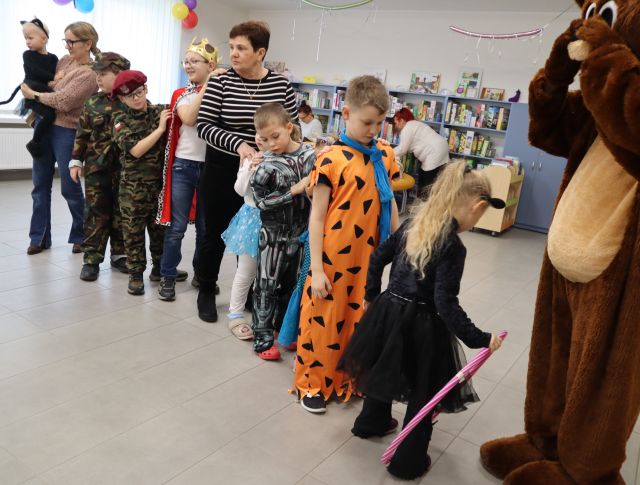 Na zdjęciu znajduje się miś, a za nim w szeregu stoją dzieci w przebraniach i dwójka dorosłych. W tle znajdują się półki z książkami.