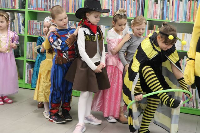 Na zdjęciu znajduje się ośmioro dzieci stojących w szeregu, pierwsza dziewczynka przekłada przez siebie hulahop. W tle znajdują się półki z książkami.