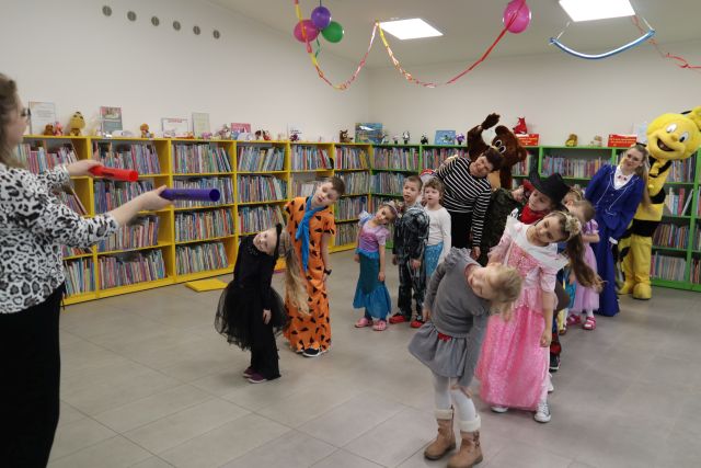 Na zdjęciu znajdują się dzieci wraz z dorosłymi w dwóch szeregach i przechylają się w bok. Przed nimi stoi kobieta, któłra rurkami pokazuje jaki mają wykonywać ruch. W tle znajdują się półki z książkami.