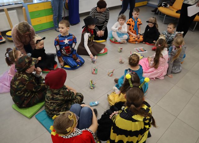 Na zdjęciu znajduje sie siedemnaścioro dzieci siedzących na podłodze, mają na sobie przebrenia, koło nich znajduja się dwie kobiety, a w środku kółka znajduja się pojemniczki z kredkami.