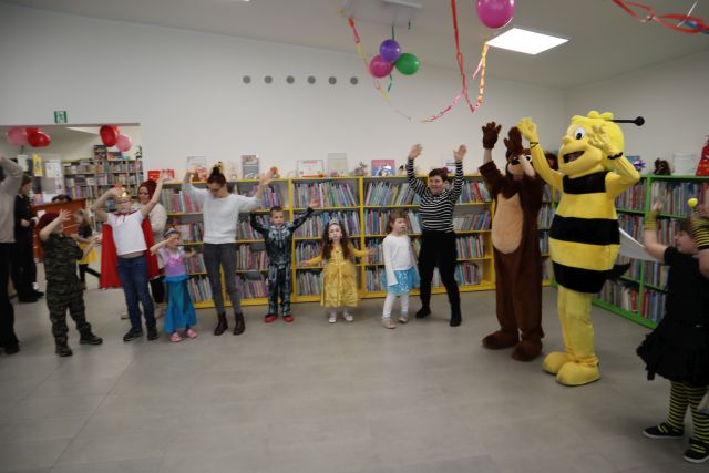 Na zdjęciu znajduję się grupa dzieci obok siebie wraz z dwójką dorosłych oraz misiem i pszczółką po prawej stronie. W tle znajdują się półki z książkami, do sufitu przywieszone są girlandy wraz z balonami.