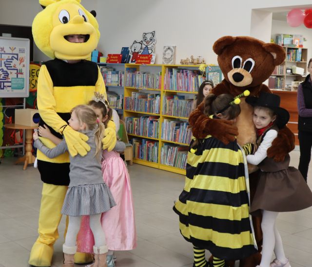 Na zdjęciu znajduje się pszółka oraz miś do których przytulają się przebrane dzieci. W tle znajdują się półki z książkami.
