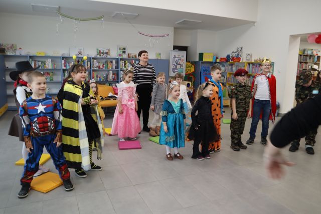 Na zdjęciu znajdują się stojące, przebrane w bajkowe stroje dzieci oraz jedna kobieta. W tle znajdują się półki z grami oraz z książkami. Po prawej stronie zdjęcia znajduje się rozmazana ręka w ruchu.