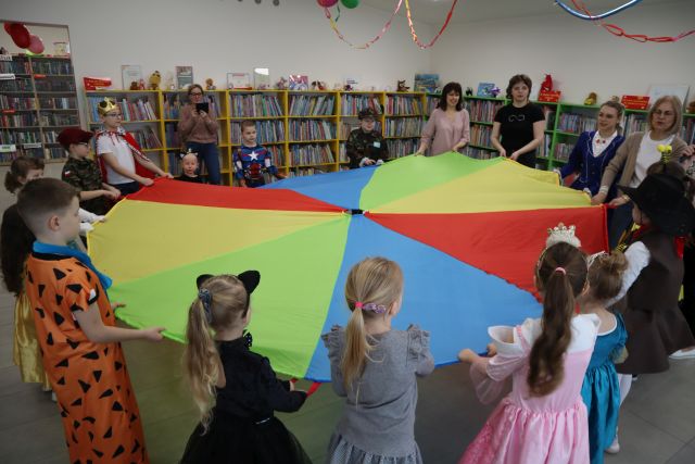 Na zdjęciu znajdują się dzieci oraz piątka dorosłych kobiet, jedna z nich robi zdjęcie telefonem, a reszta wraz z dziećmi trzyma kolorową okragłą chustę. W tle znajdują się półki z książkami, a z sufitu zwisają kolorowe girlandy.