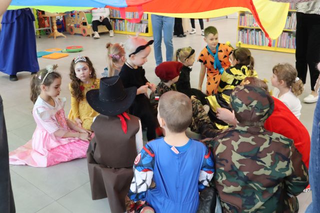 Na zdjęciu znajdują się dzieci na podłodze, a nad nimi unosi się kolorowa chusta trzymana przez kobiety. W tle znajdują się półki z książkami.