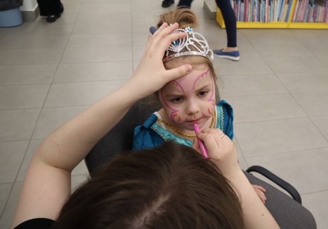 Na zdjęciu znajduje się dziewczynka, które kobieta przed nią maluje na twarzy motylka. Dziewczynka jest przebrana za księżniczkę i ma na sobie koronę. W tle znajduje się podłoga oraz półki z książkami.